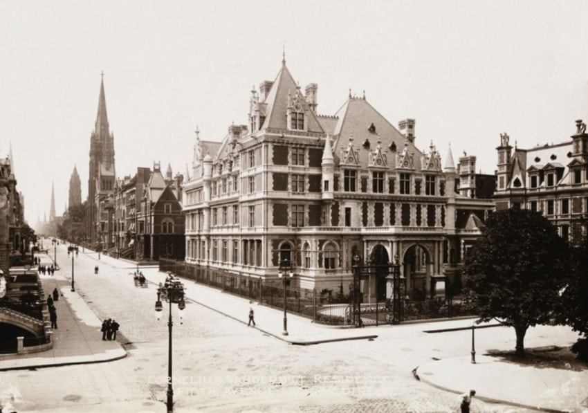 Historic city street with ornate buildings and a tall church spire, pedestrians walking along the sidewalks, early 1900s.