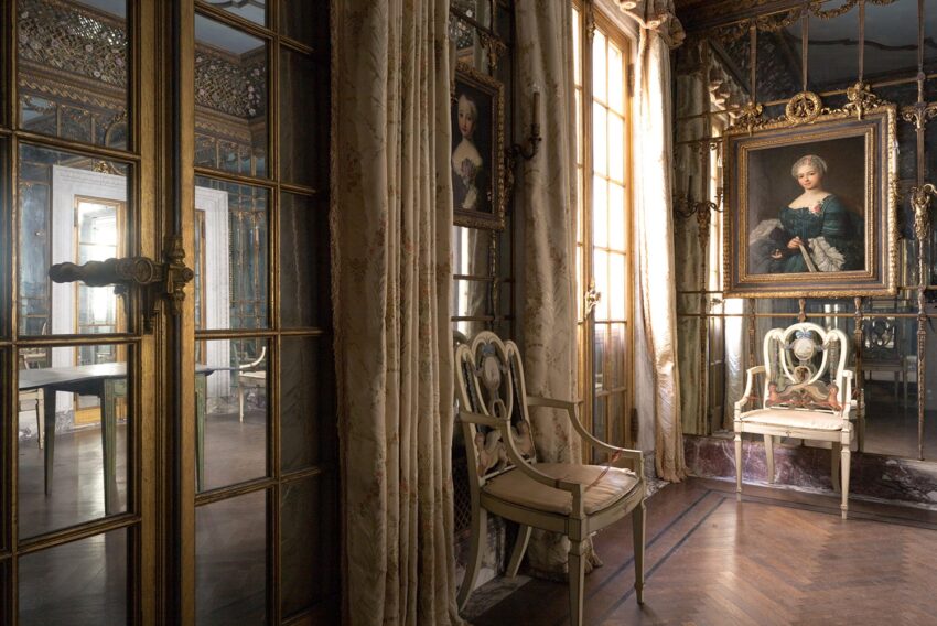 Elegant vintage room with ornate mirrors, framed portraits, wooden chairs, and tall windows draped with floral curtains.