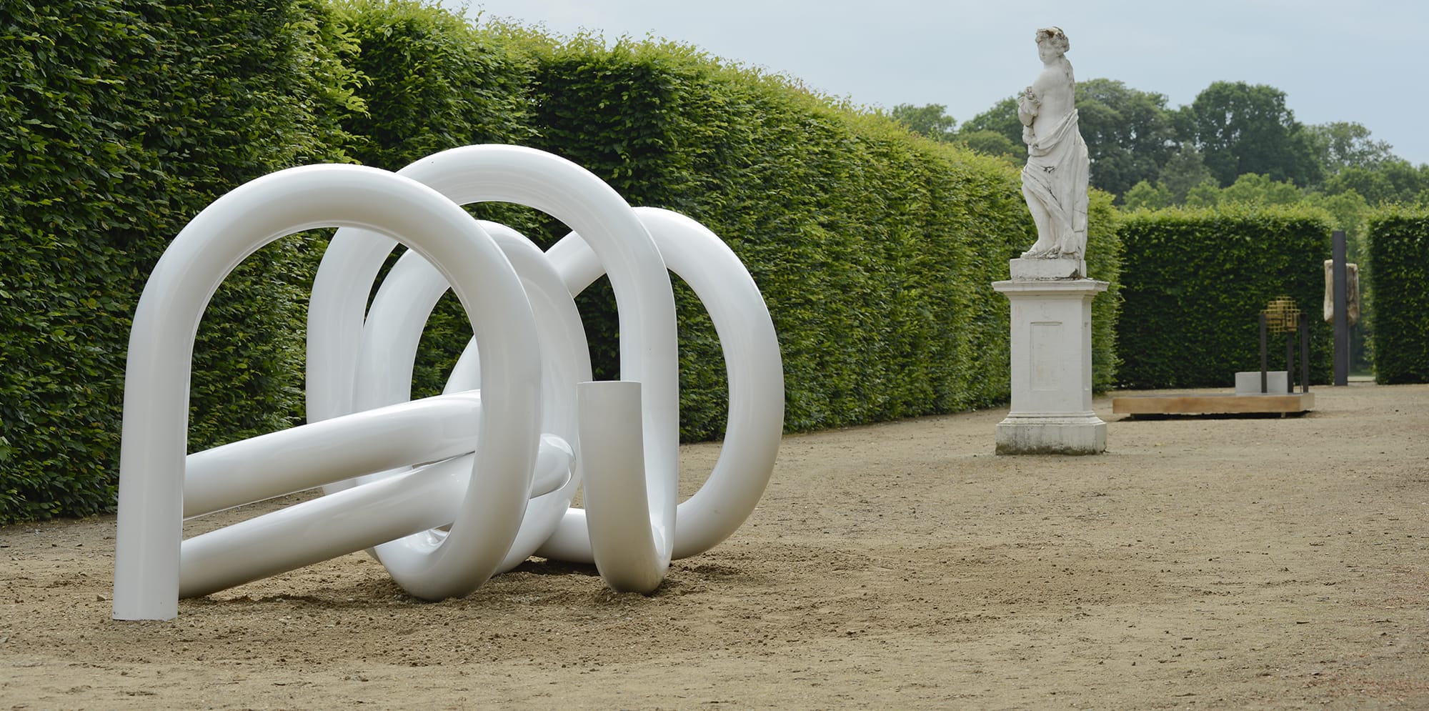 White abstract sculpture in a garden with a classical statue and manicured hedges in the background.