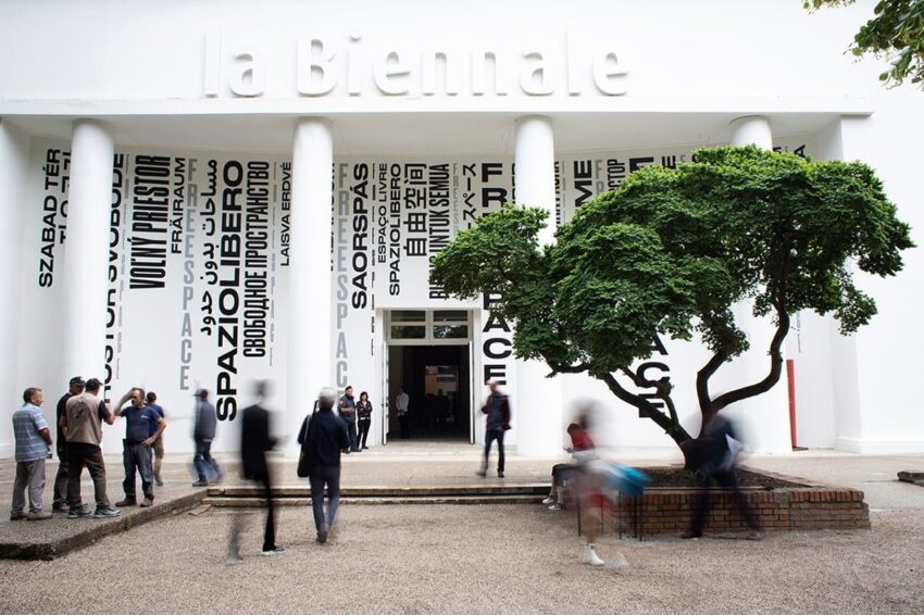 People walking outside La Biennale building with modern art posters and a lush green tree in the foreground.