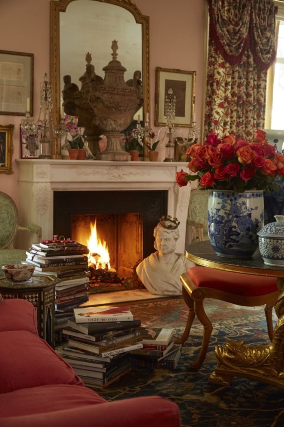 Cozy living room with a lit fireplace, ornate bust, stacks of books, and vase of red roses on a colorful rug.