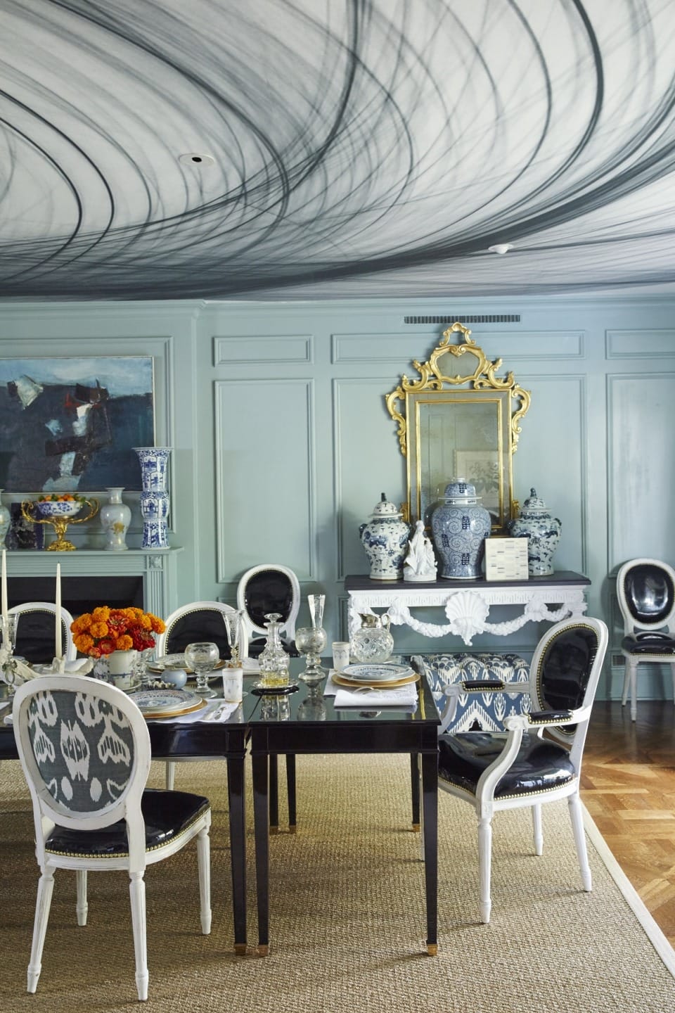 Elegant dining room with ornate chairs, dark table, decorative vases, large mirror, artwork, and a unique swirling ceiling design.