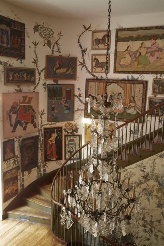 Spiral staircase with ornate chandelier and walls adorned with various framed art and floral wallpaper.