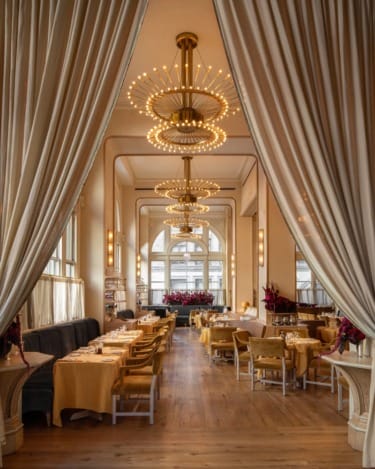 Elegant restaurant interior with chandeliers, draped curtains, wooden flooring, and tables set for dining.