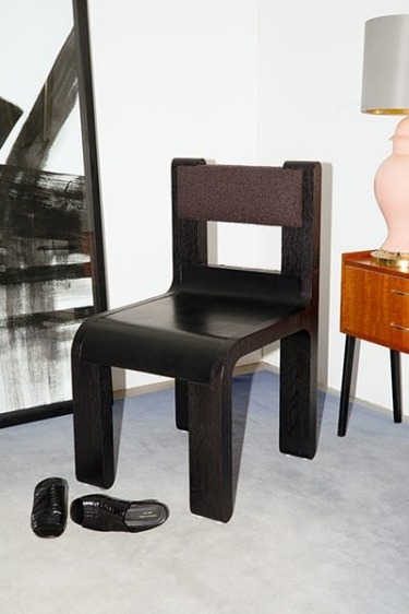 Modern black wooden chair in a room with abstract art, a wooden cabinet with a pink lamp, and a pair of black slippers.