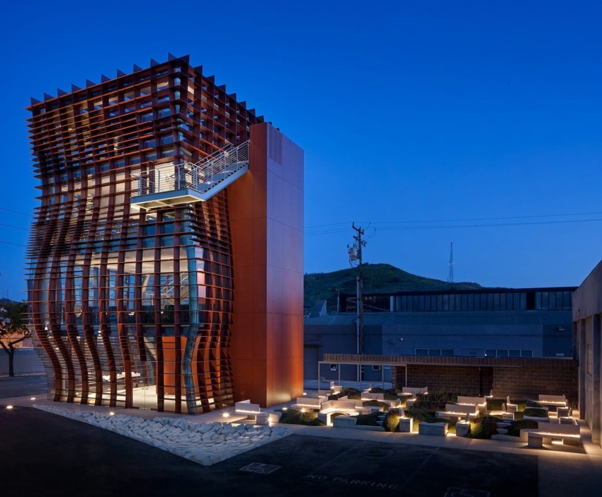 Modern building with a unique wavy facade pattern, lit up at night, surrounded by a landscaped area and nearby urban structures.