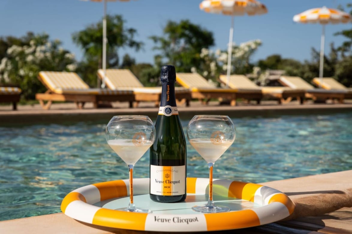 Tray with a Veuve Clicquot champagne bottle and two glasses by a poolside with lounge chairs and umbrellas in the background.