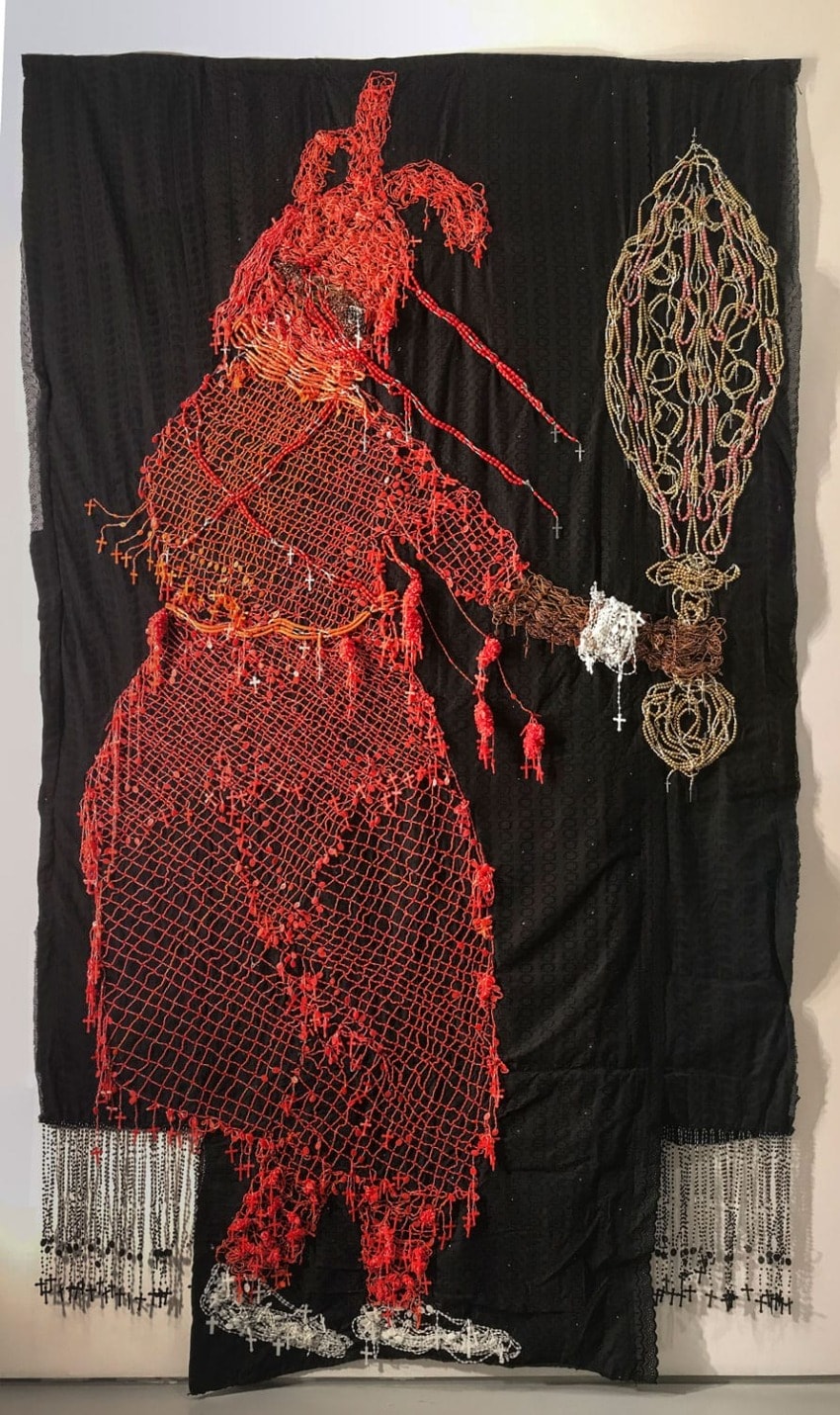 Textile artwork of a large red figure holding a woven, elongated object on a black background with intricate patterns.