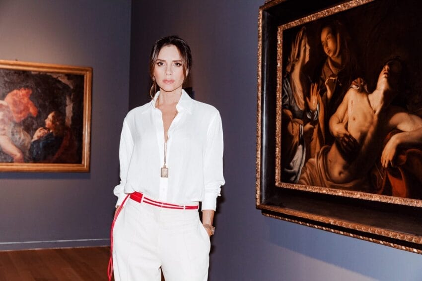 Person in white outfit stands in an art gallery with classical paintings in the background.