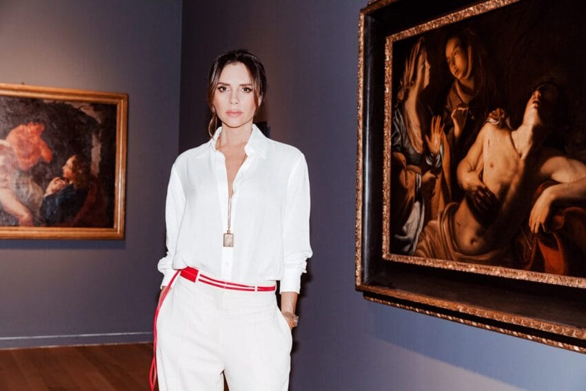 Person in white outfit stands in an art gallery with classical paintings in the background.