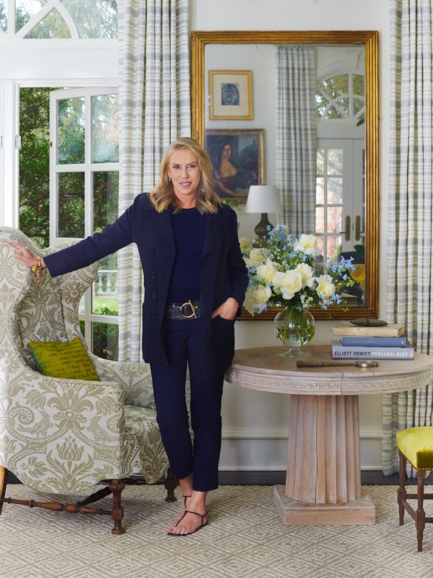 Person in a navy suit stands by armchair in a elegantly decorated room with flowers, mirror, and large windows.