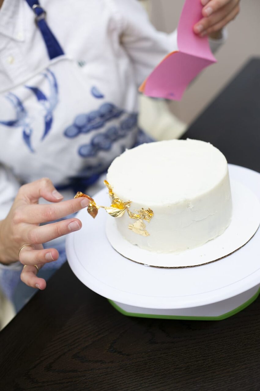 Person applying gold leaf to a white frosted cake on a rotating stand.