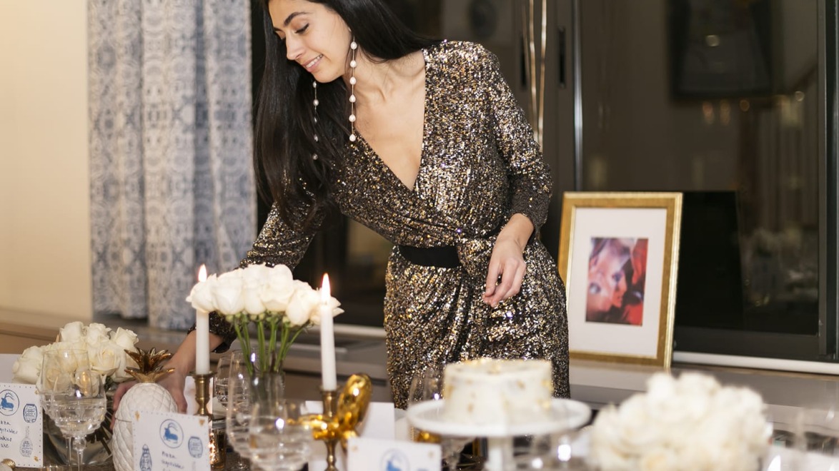 Woman setting a decorated dinner table with candles and flowers, wearing a sparkling dress in a festive setting.