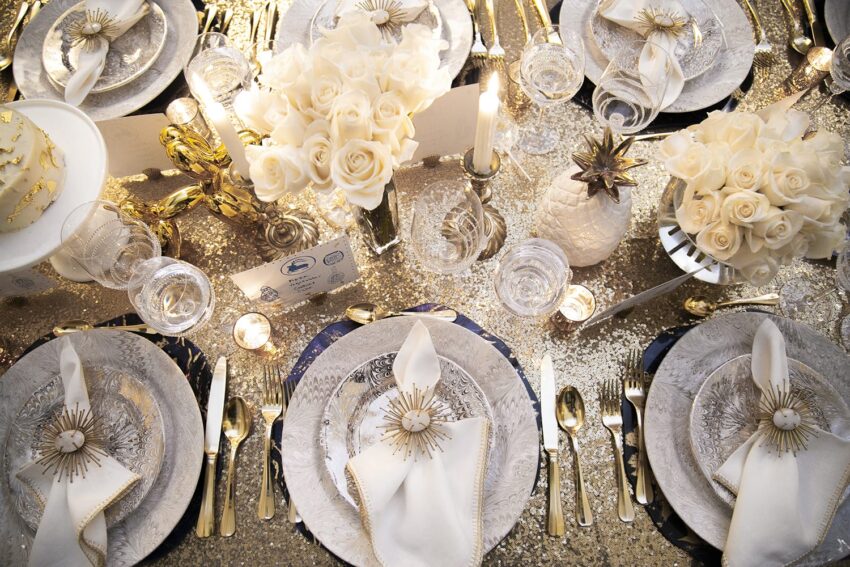 Elegant table setting with golden cutlery, white roses, and decorative pineapples on a sparkling sequin tablecloth.
