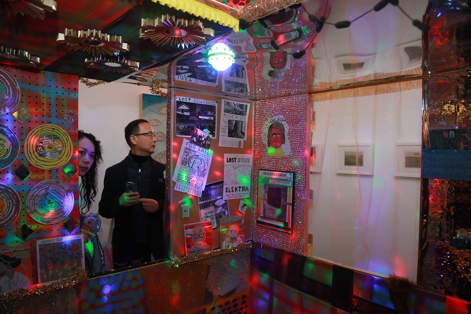 Two people observing a vibrant, eclectic art installation with colorful lights, posters, and reflective surfaces.
