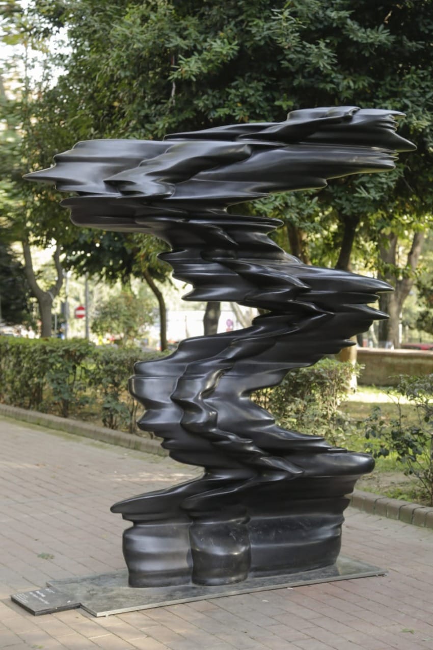 Abstract black sculpture with twisted, wavy shapes, set outdoors on a paved path surrounded by trees and greenery.