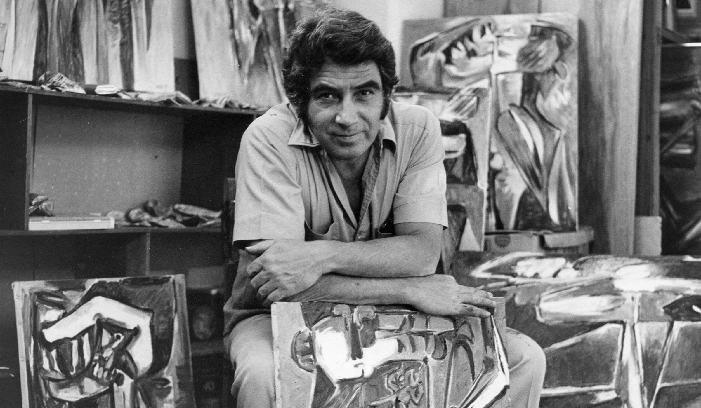Artist sitting among abstract paintings in a studio, with various artworks surrounding him.
