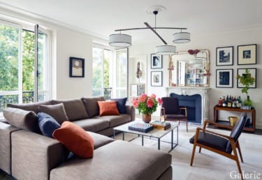 Modern living room with large gray sectional, colorful cushions, artwork on walls, and bright natural light from windows.