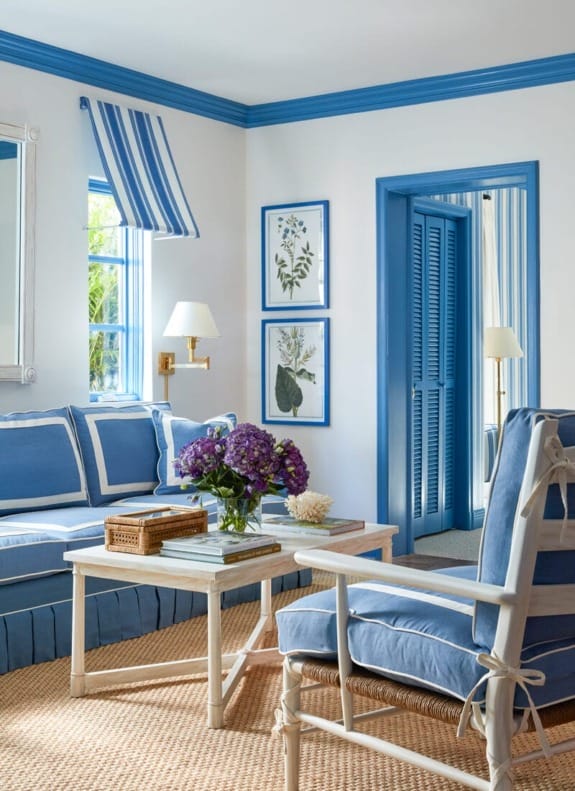Blue and white living room with a striped couch, armchair, floral artwork, and a wooden coffee table with purple flowers.