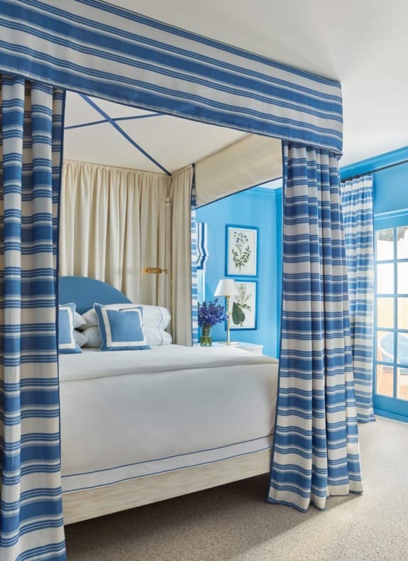 Four-poster bed with blue and white striped curtains, decorated with cushions, flowers, and framed botanical prints on the walls.