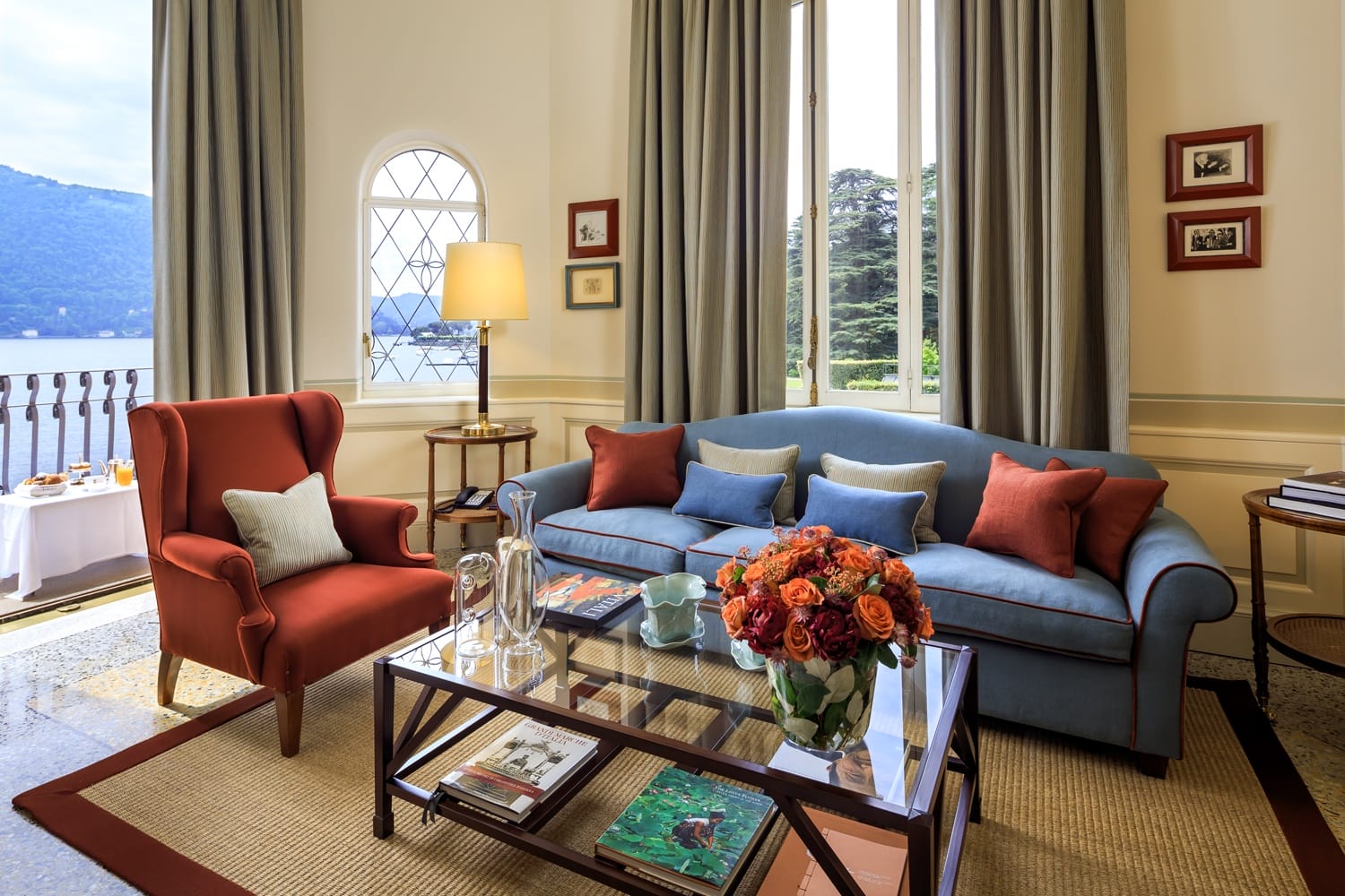 Elegant living room with blue sofa, red armchair, glass coffee table, floral centerpiece, and large windows overlooking a lake.