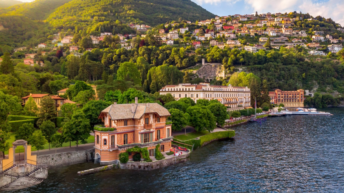 Aerial view of colorful houses and greenery near a calm lake with hills in the background under a partly cloudy sky.