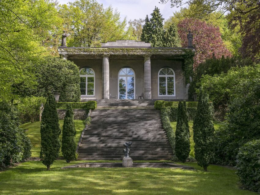 Small stone building with large windows and columns, surrounded by lush greenery and a manicured garden with steps in front.