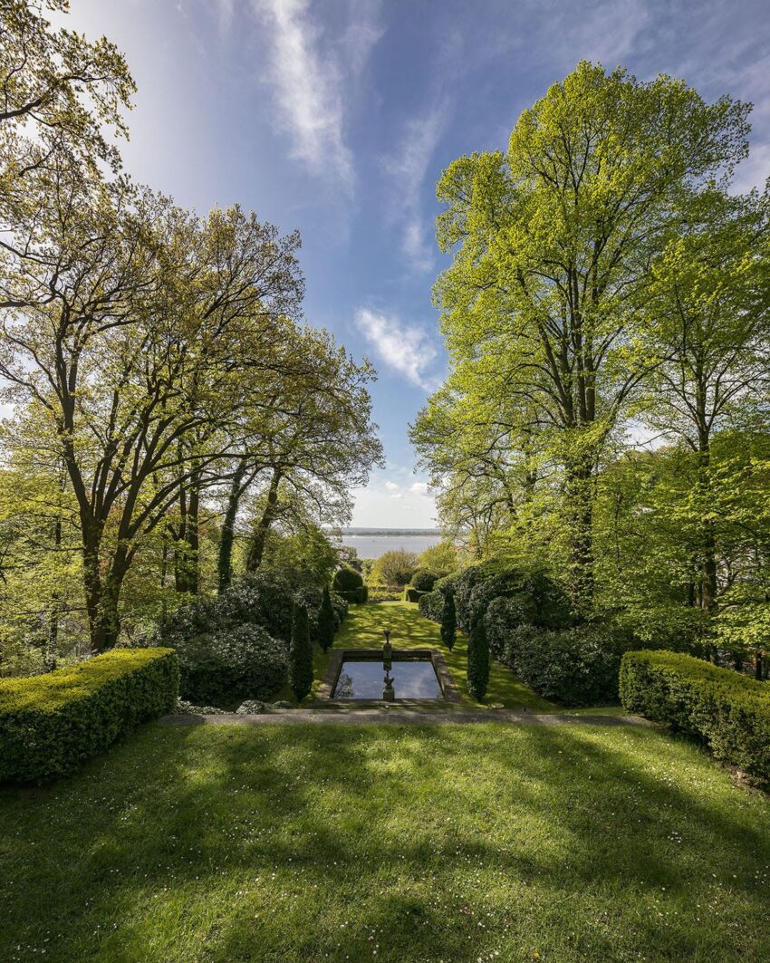 Scenic garden view with lush green trees, manicured hedges, and a pathway leading to a water feature under a blue sky.