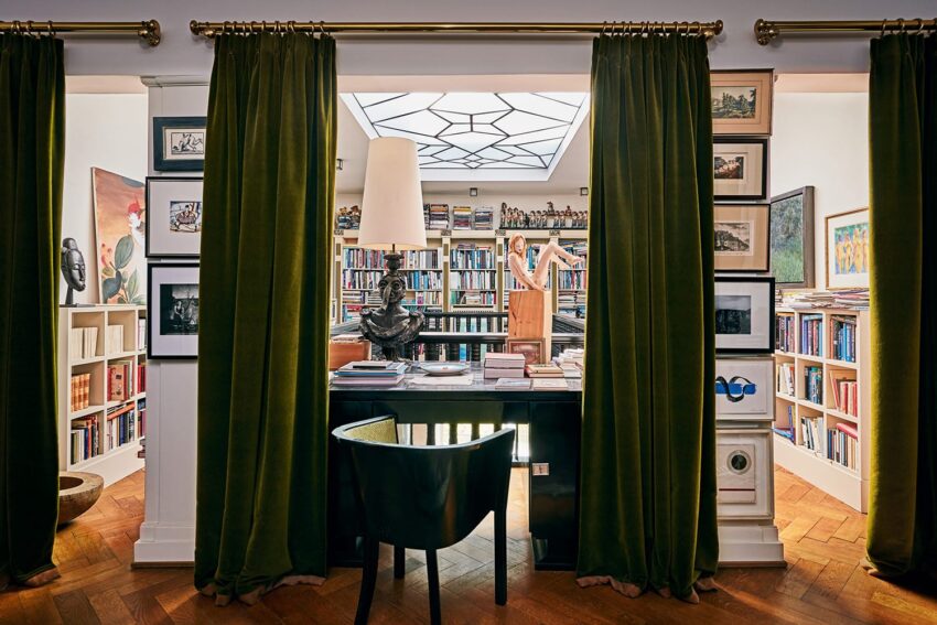 Elegant study with green curtains, a desk, and a chair, surrounded by bookshelves filled with various books and decorations.