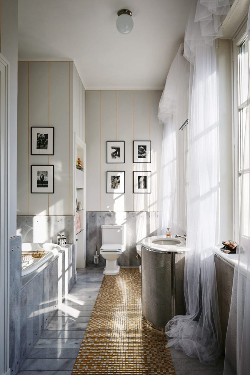 Bright bathroom with framed pictures, bathtub, toilet, and a gold-tiled floor. Sunlight streams through sheer curtains.