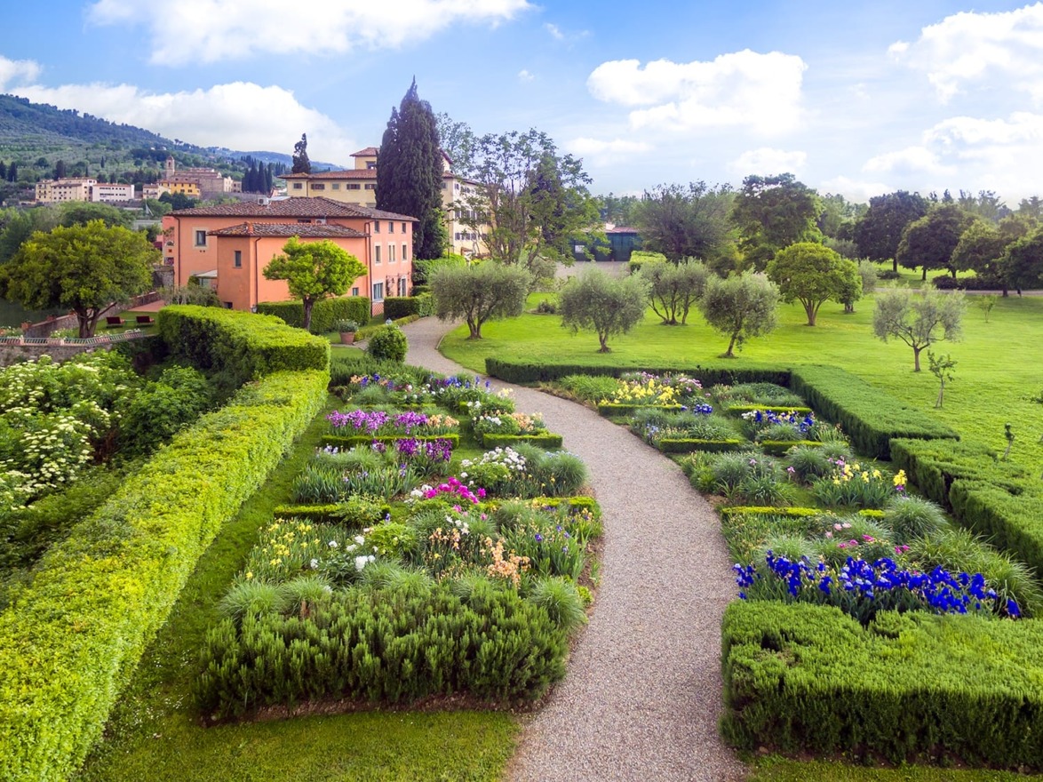 Aerial view of a colorful garden with a winding path, surrounded by trees and a villa in the background on a sunny day.