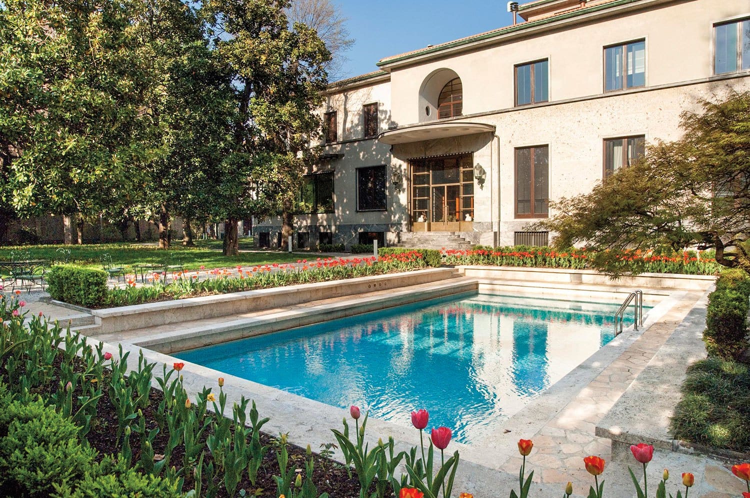 Elegant villa exterior with a rectangular swimming pool surrounded by blooming tulips and greenery on a sunny day.