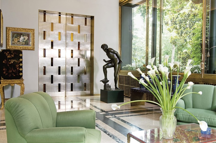 Elegant living room with green chairs, a vase of white flowers, a bronze sculpture, large windows, and a modern metallic door.