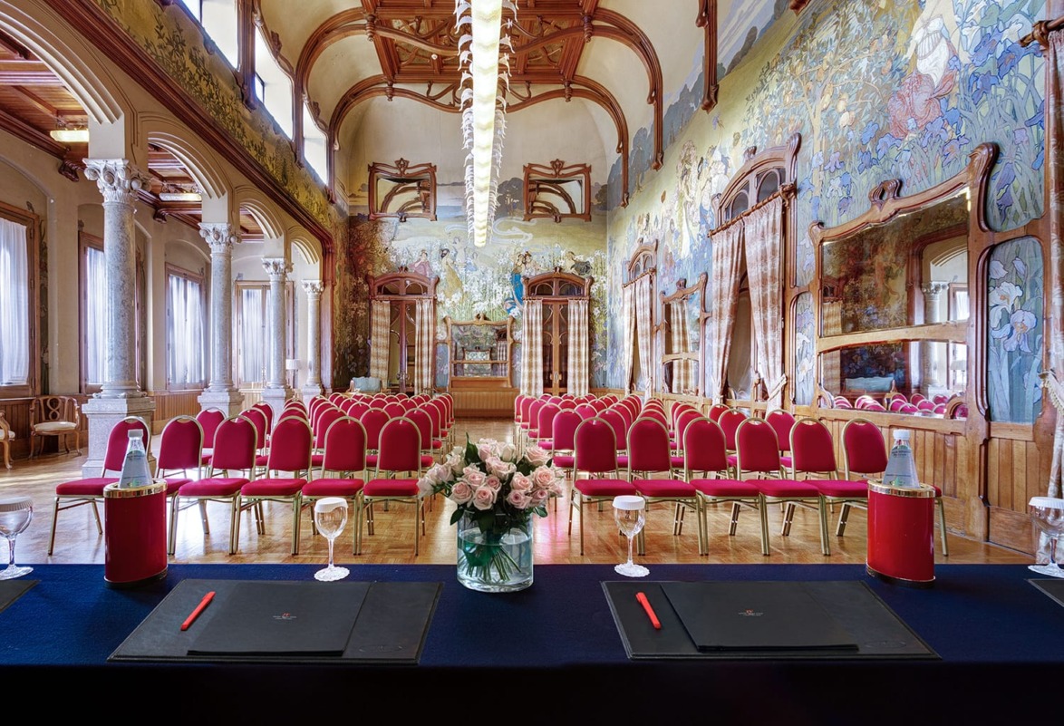 Ornate conference room with mural walls, red chairs, and a table set with flower bouquets, water bottles, and notepads.