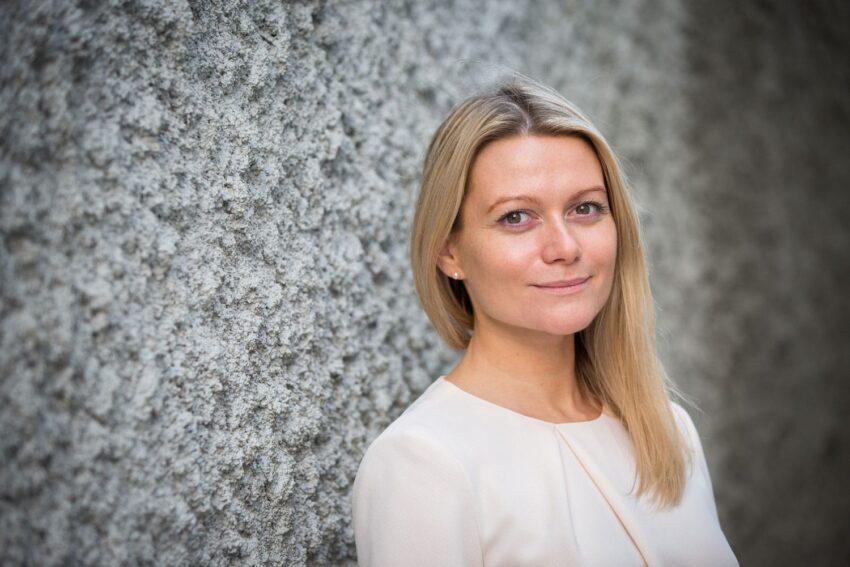 Person with long blonde hair standing against a textured grey wall, wearing a light-colored top and smiling softly.
