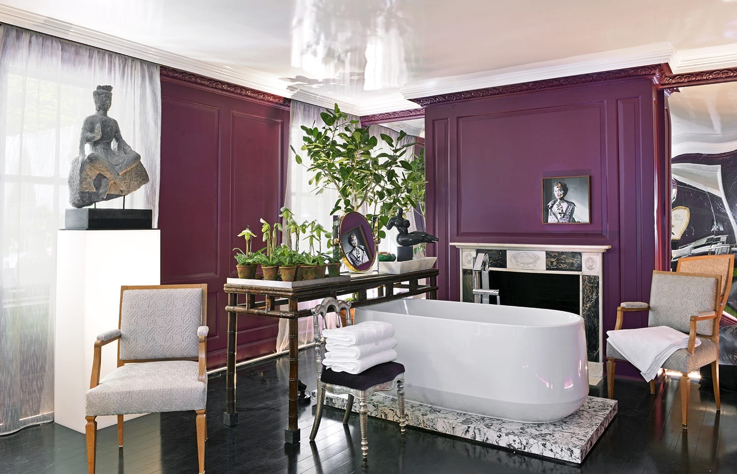 Luxurious bathroom with freestanding tub, purple walls, greenery, sculptures, and modern furniture.