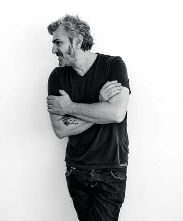 Man with beard and casual attire standing against a white background, arms crossed and looking to the side. Black and white photo.