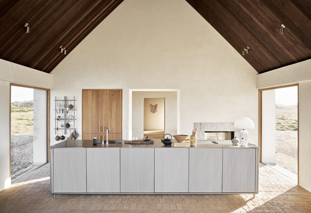 Modern kitchen interior with minimalist design, large island, vaulted wooden ceiling, and natural light from open doors.