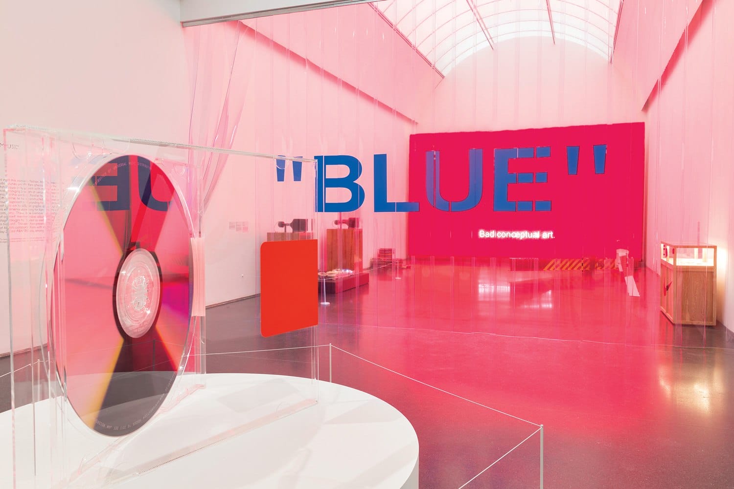 Art installation with a large transparent disc and the word "Blue" on a pink-lit background in a modern gallery setting.