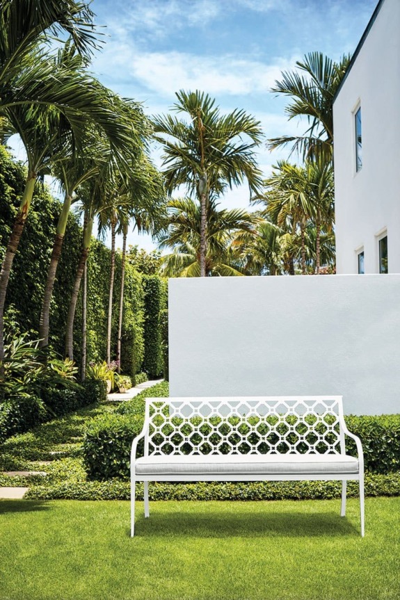White metal bench on green lawn with palm trees and modern white building in the background on a sunny day.