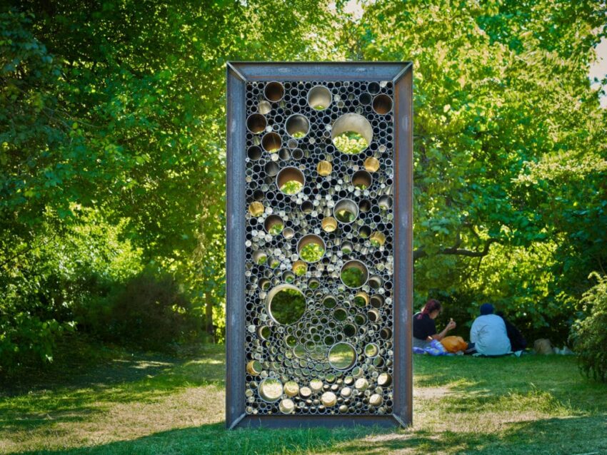 Metal sculpture with circular patterns standing on grass in a park with people sitting in the background and trees around.