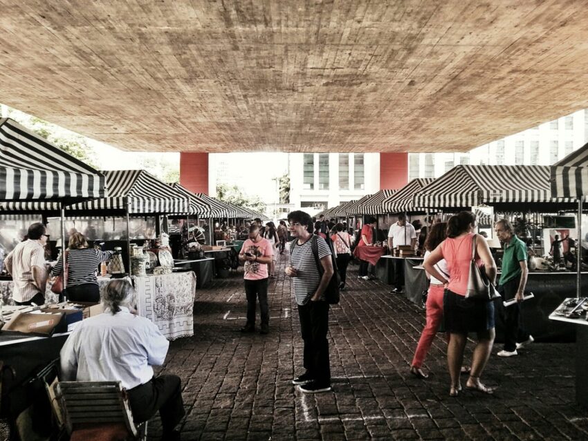 People browsing a bustling outdoor market with striped tents under a large concrete structure on a sunny day.