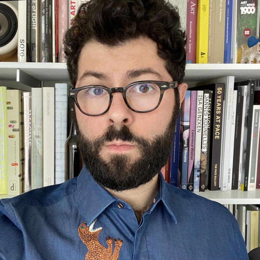 A person with glasses and a beard stands in front of a bookshelf wearing a blue shirt with an embroidered bear design.