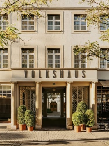 Facade of a beige building with "Volkshaus" sign, framed by trees and potted shrubs, under a clear sky.