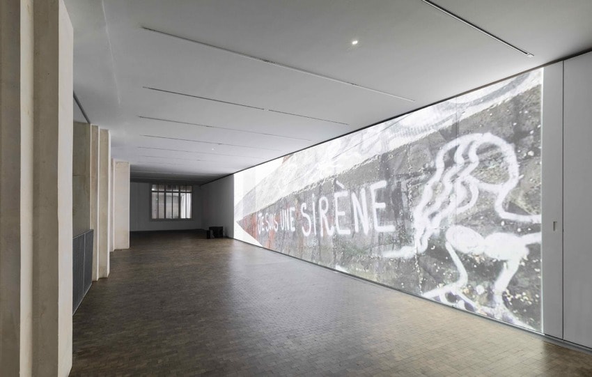 Art installation with projected graffiti of mermaid and text in minimalist gallery space with tiled floor and high ceiling.
