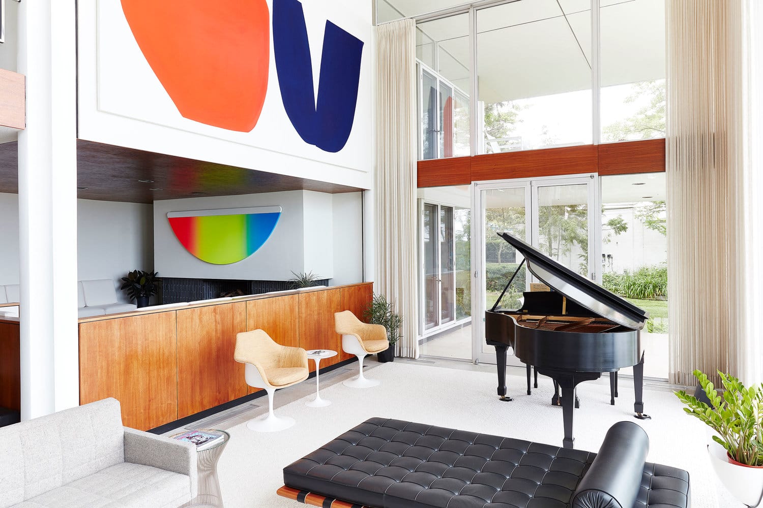 Modern living room with grand piano, colorful abstract art on walls, white chairs, wooden accents, and large windows.