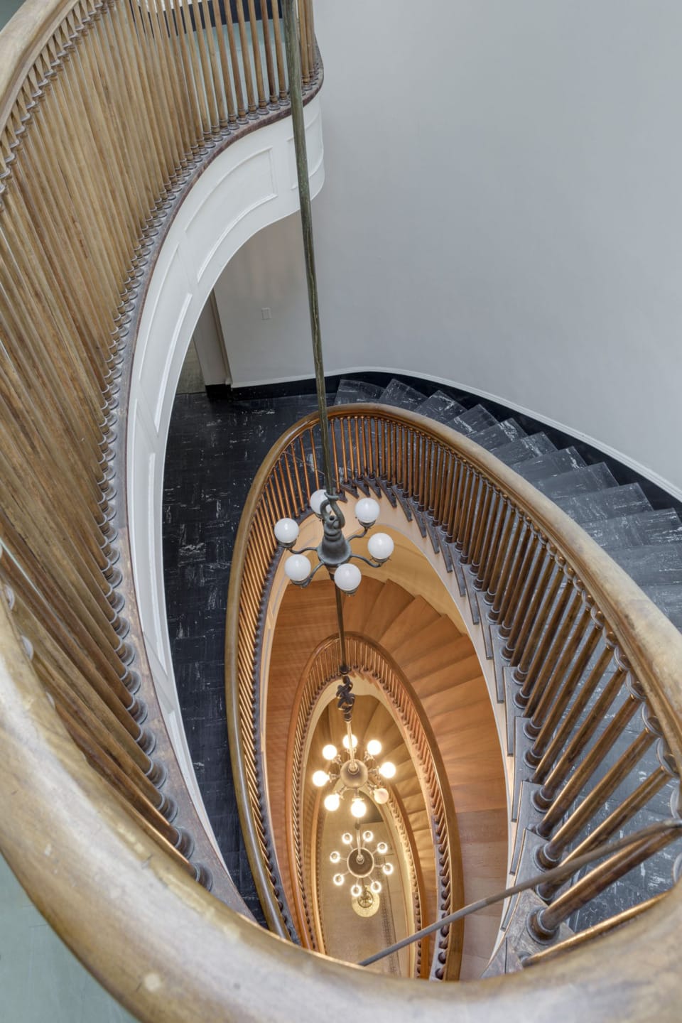 Spiral staircase with wooden railings and hanging lights descending to a dark floor.