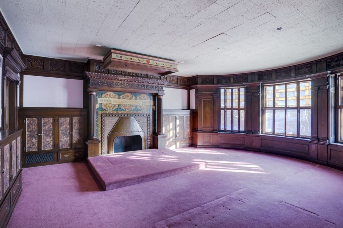 Room with purple carpet, ornate wooden fireplace, and large bay windows allowing natural light to fill the space.