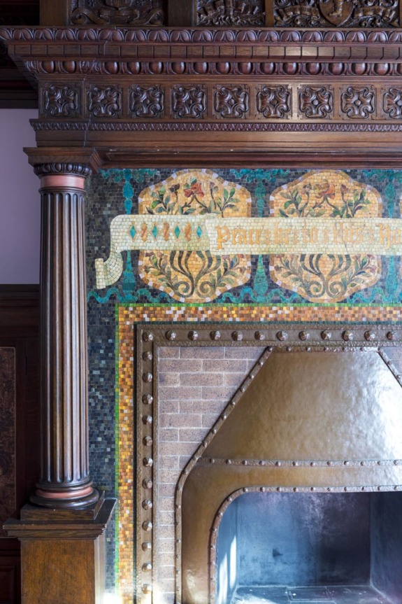 Ornate wooden fireplace with intricate carvings, colorful mosaic tiles, and decorative columns in a historical setting