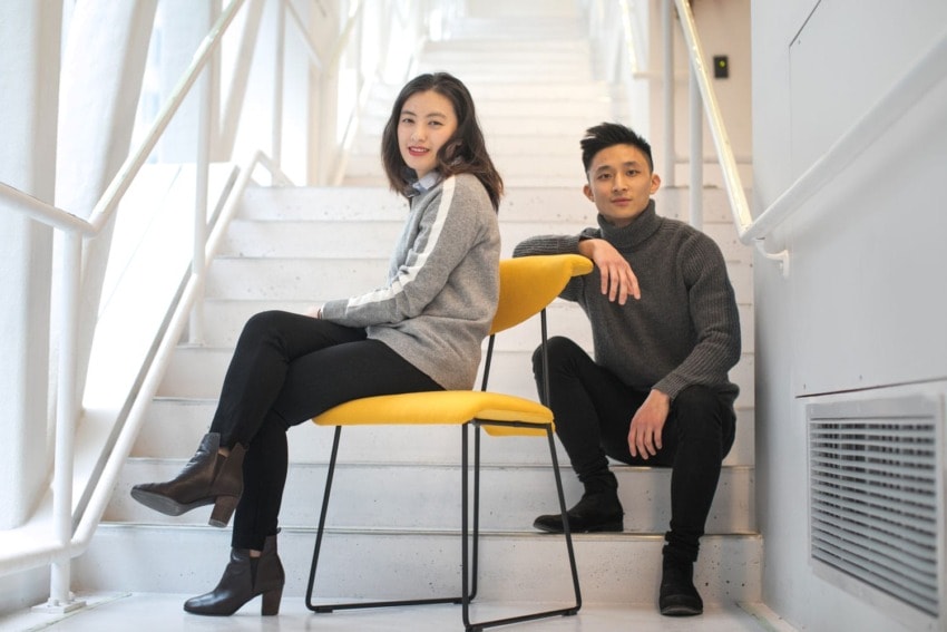 Two people posing on a staircase, one seated on a yellow chair and the other sitting on a step, both in casual clothing.